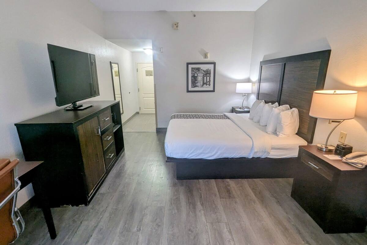 Une chambre d’hôtel avec un lit, une télévision, une commode, une table de chevet avec une lampe et un tableau encadré au mur.