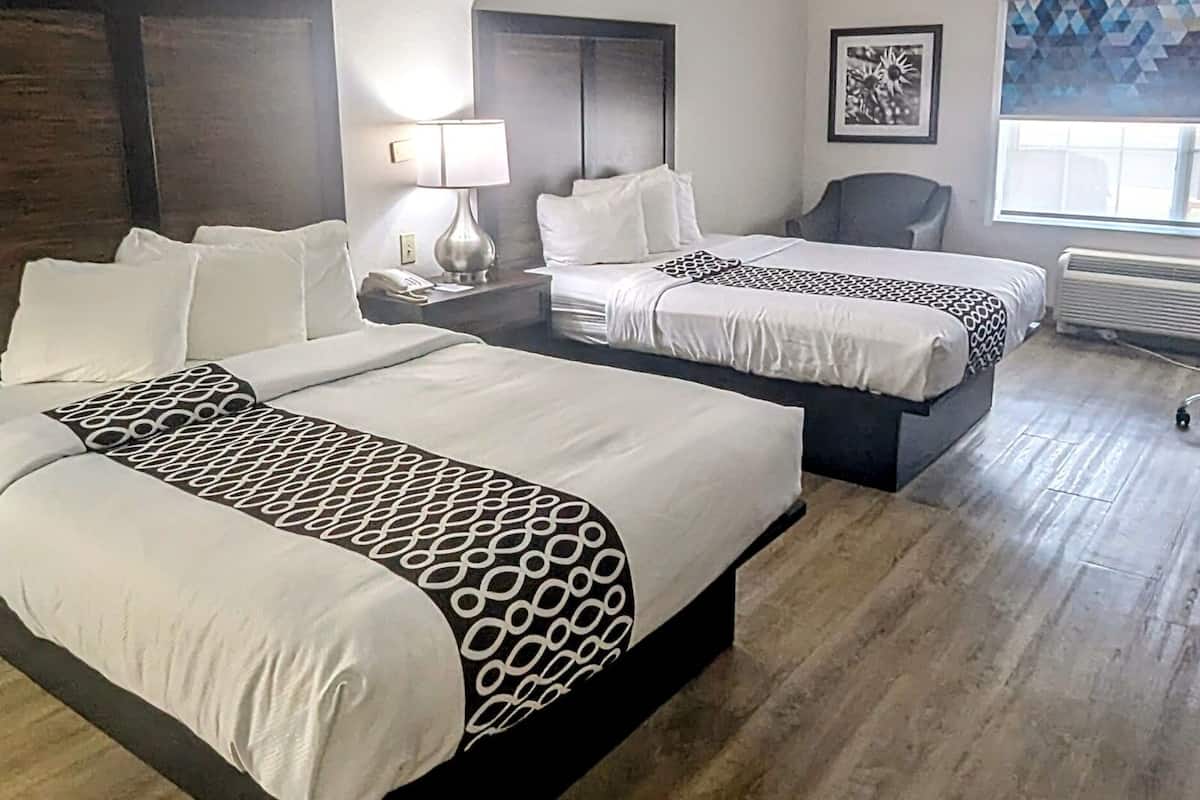 Une chambre d’hôtel avec deux lits, une chaise, une lampe, un tableau encadré et une fenêtre avec un rideau à motifs géométriques.