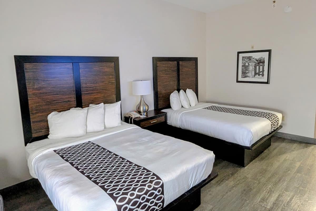 Une chambre d’hôtel avec deux lits, chacun doté d’une tête de lit en bois, de draps blancs et d’un couvre-lit à motifs. On y trouve une table de chevet avec une lampe et un tableau encadré accroché au mur.