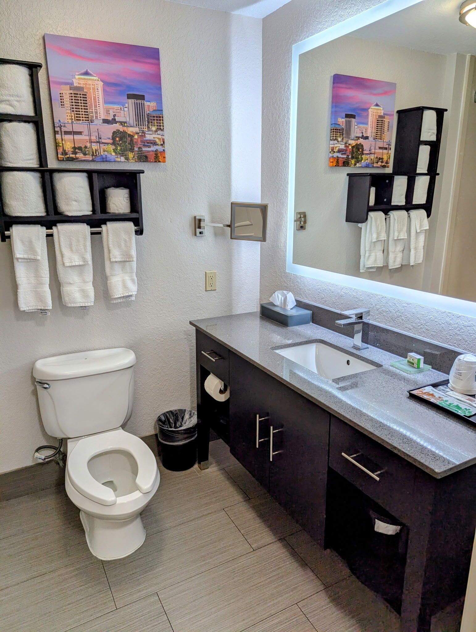 Un baño moderno con inodoro, lavabo y espejo. Hay toallas colgadas en estantes y un bote de basura.