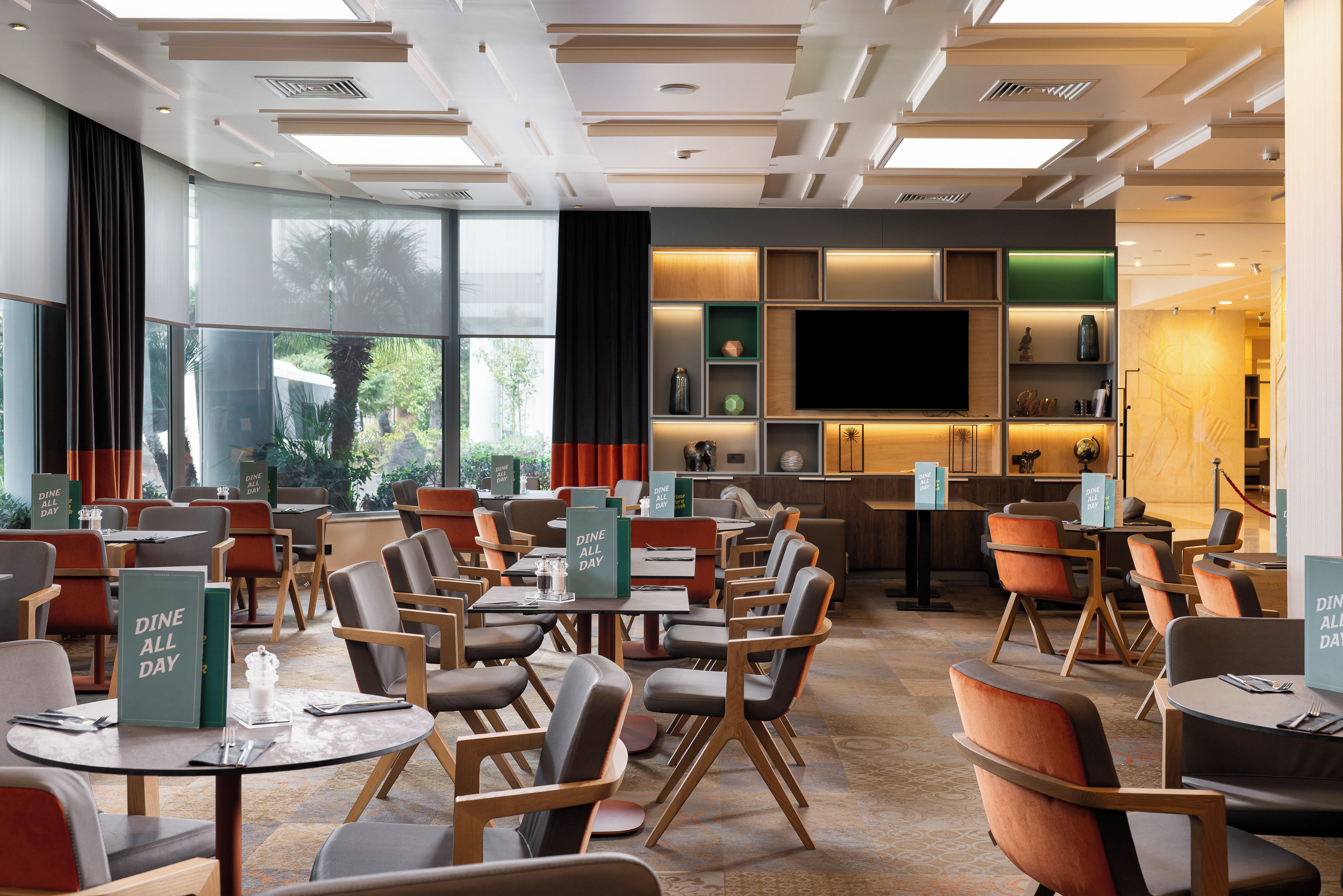 A modern hotel dining area with tables, chairs, a TV, and a view of the outdoors.