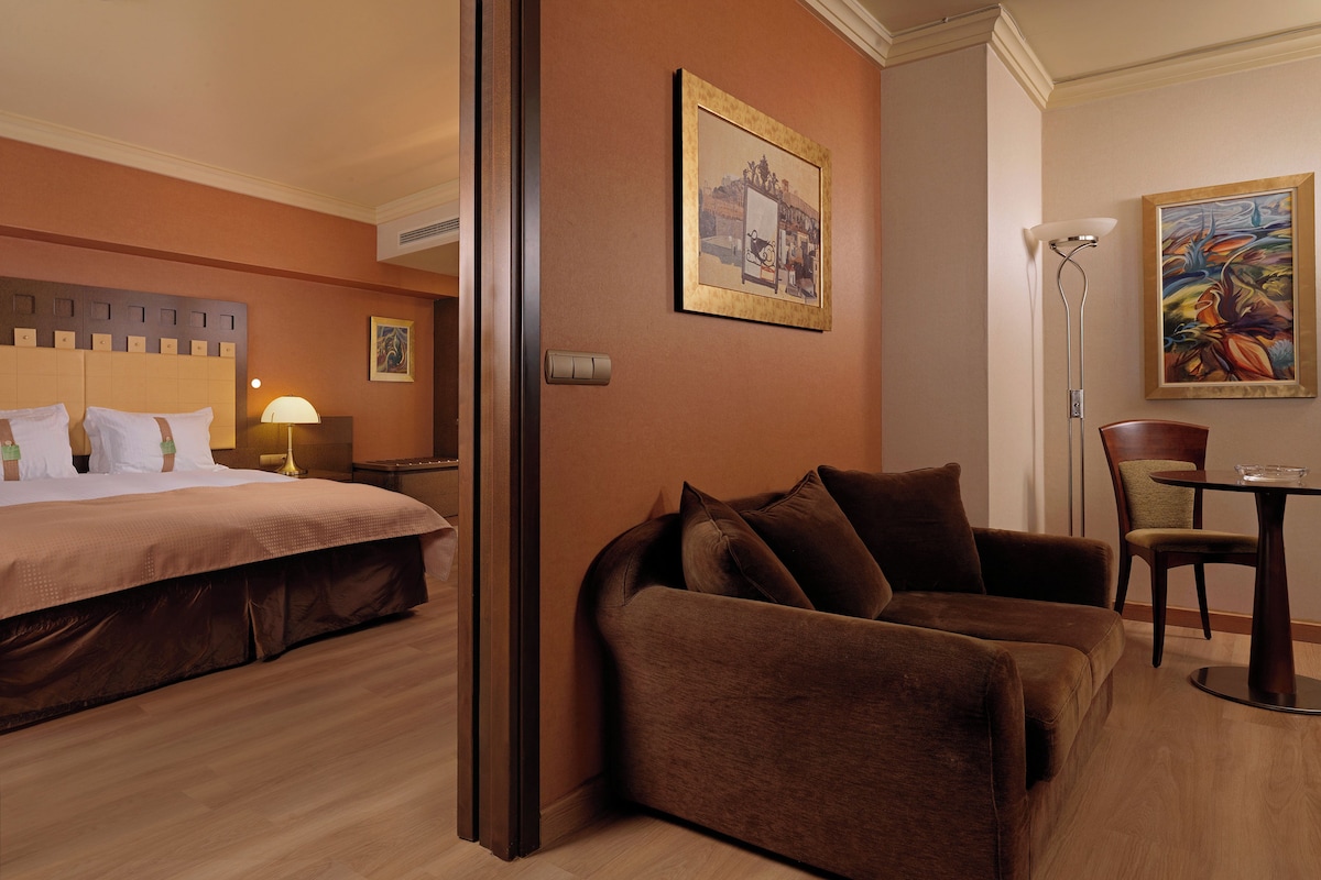 A hotel room with a bed, a brown sofa, a dining table with a chair, and two paintings on the walls.
