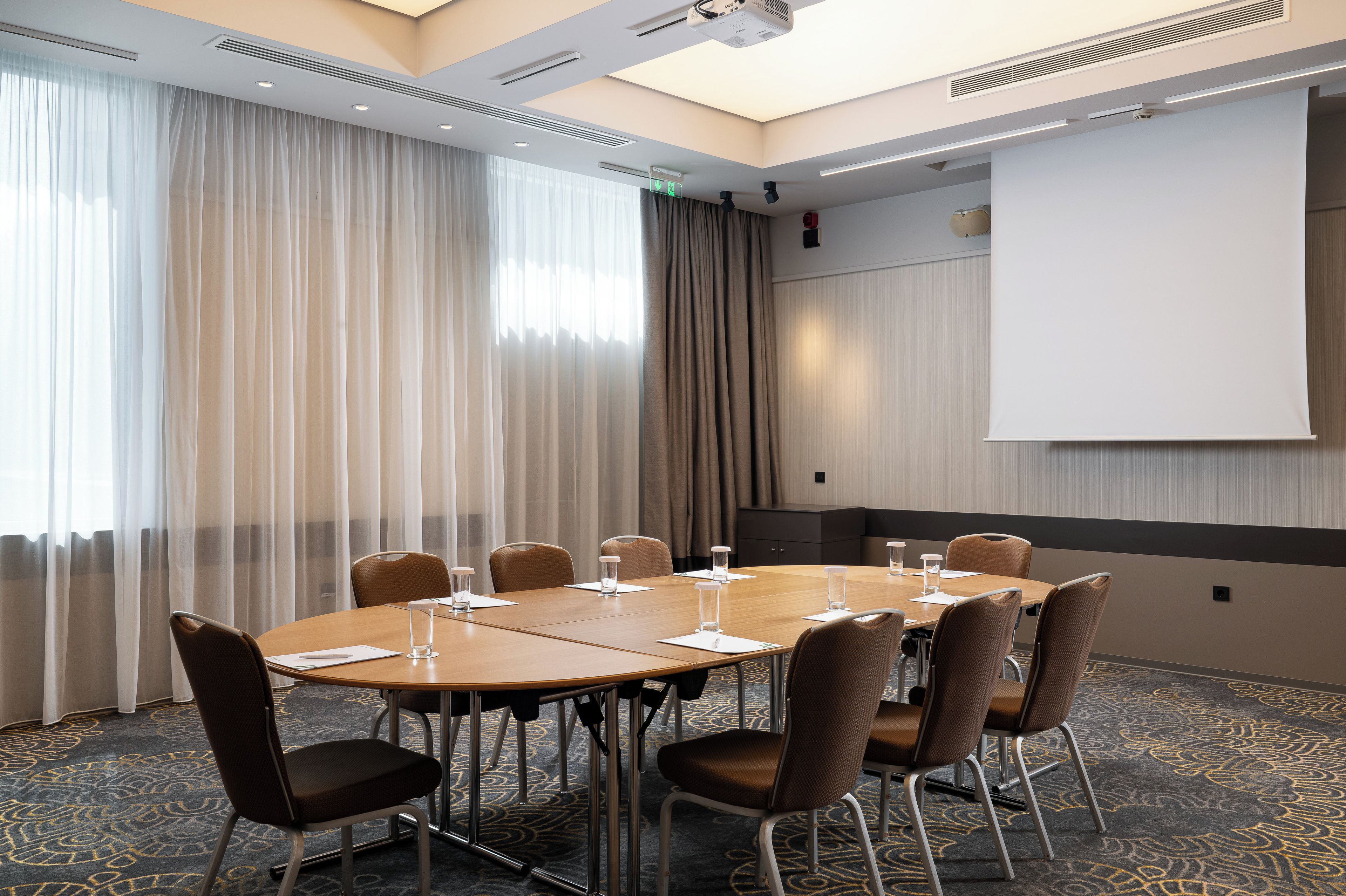 A conference room with a round table, chairs, a projector screen, and a large window with curtains.