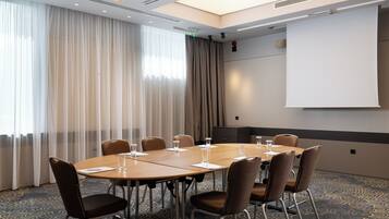 Un salón de conferencias con una mesa redonda, sillas, una pantalla de proyector y un amplio ventanal con cortinas.