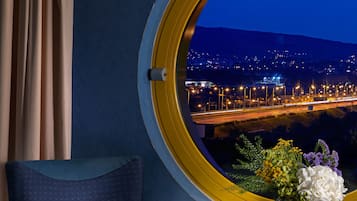 A room with a round window overlooking a cityscape at night, a blue armchair, a small table with a vase of flowers, and a curtain.