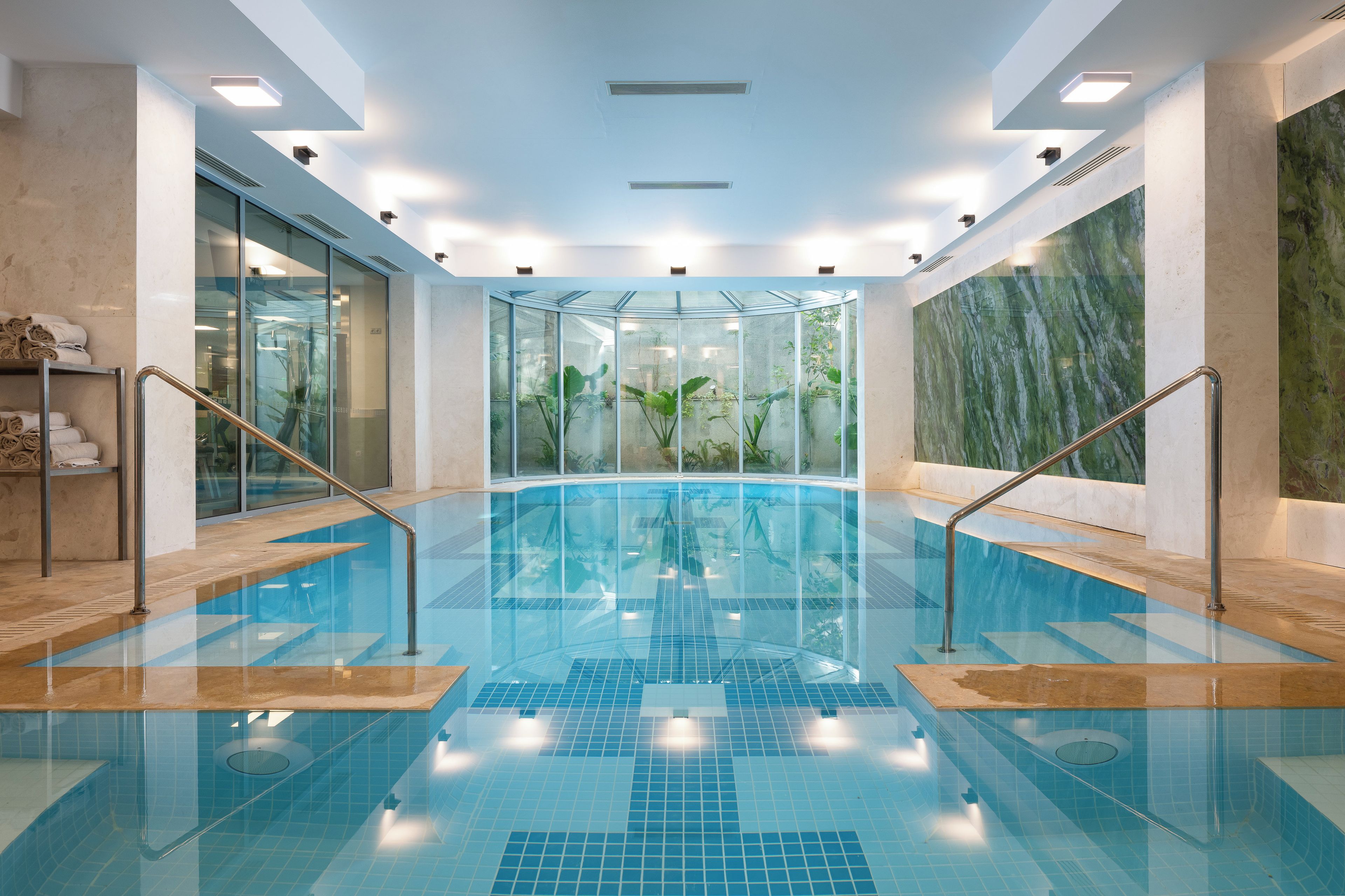 Una piscina interior con paredes de vidrio, barandales metálicos y suelo de baldosas.