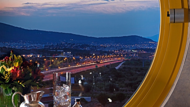 Une table avec un journal, un stylo, un pichet en verre et une bouteille, vue à travers une fenêtre donnant sur un paysage urbain au crépuscule.