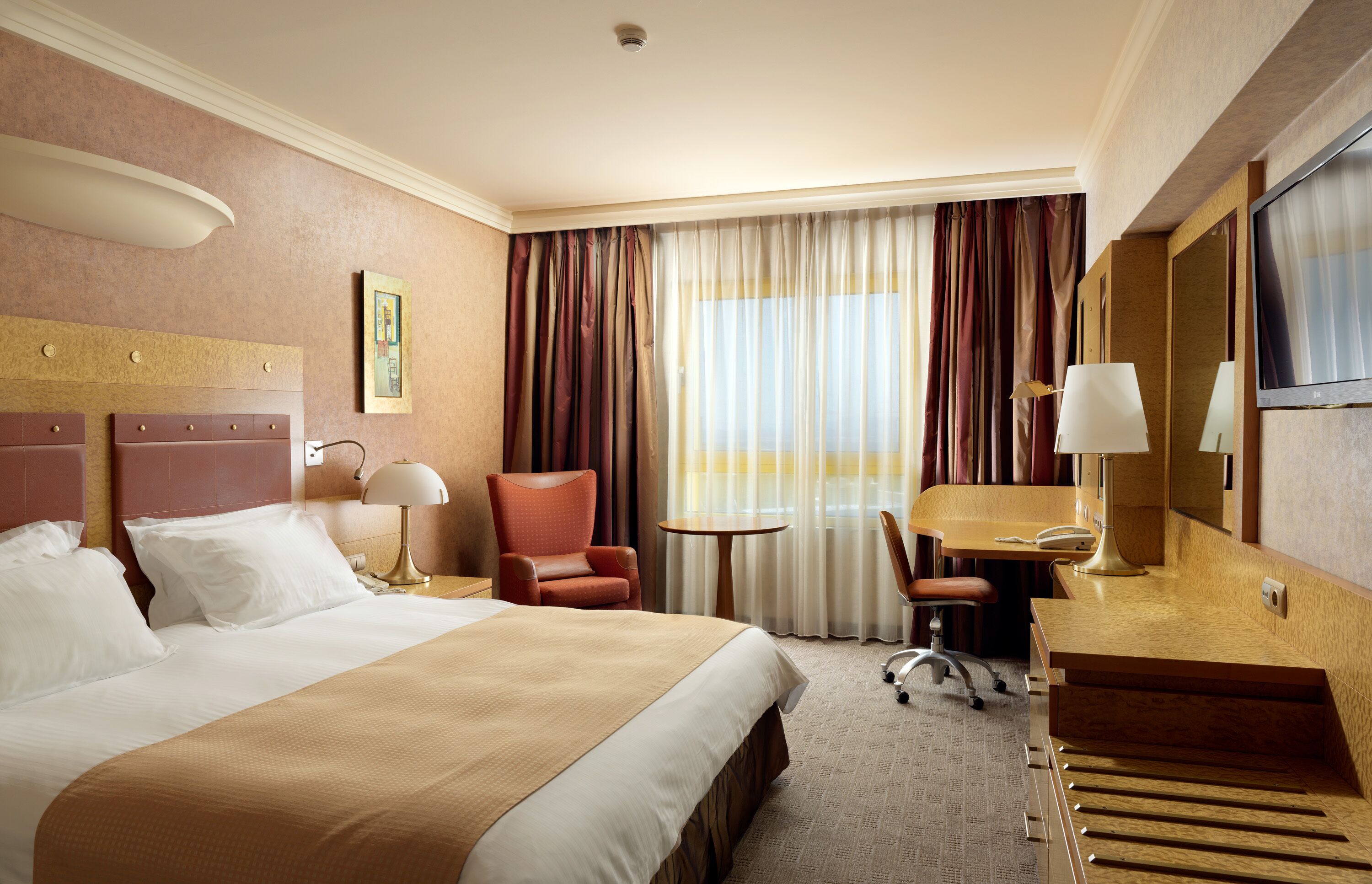 A hotel room with a large bed, a desk, a chair, a small table, a mirror, and a television.
