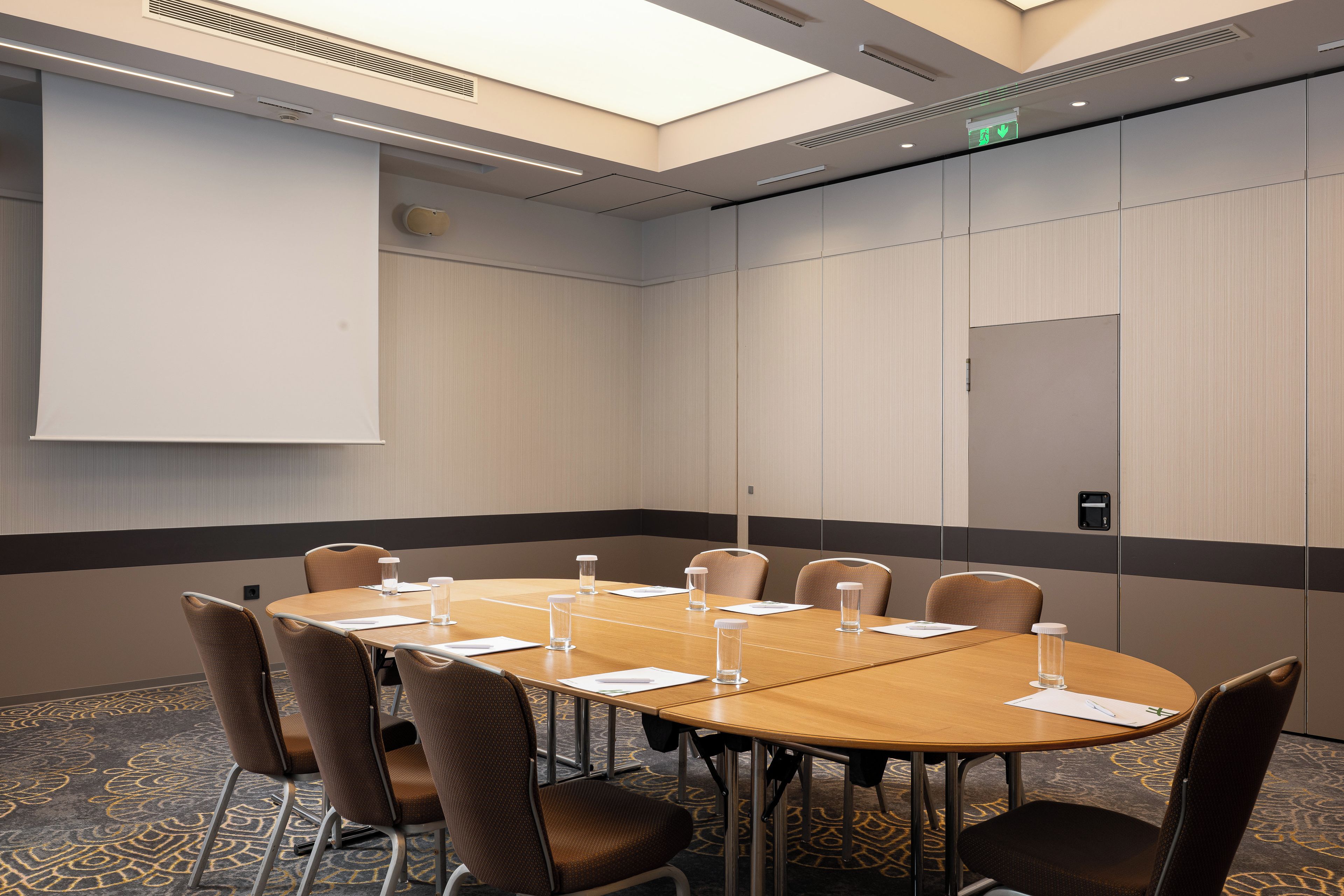 A conference room with a long table, chairs, a projector screen, and a patterned carpet.