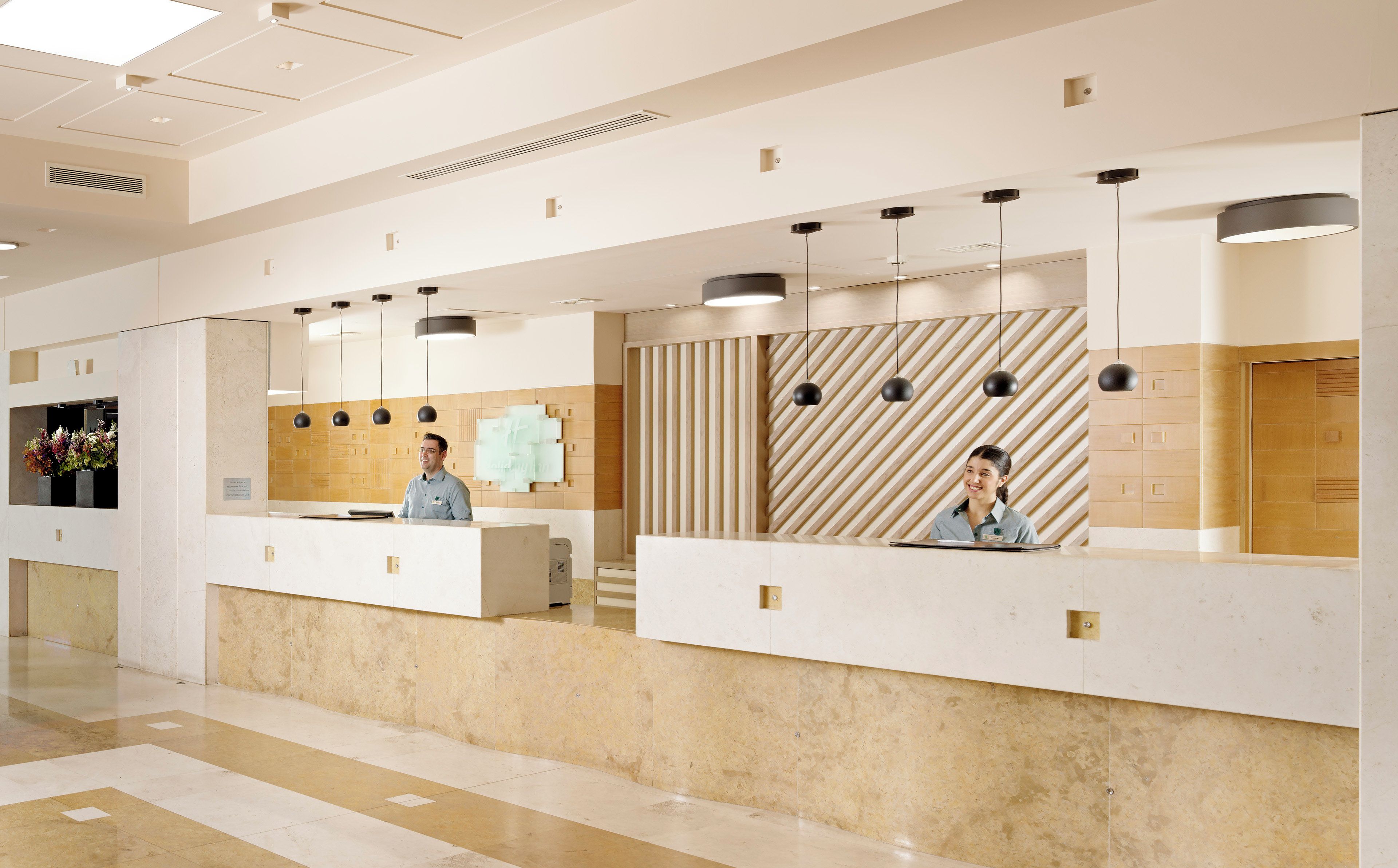 Een hotel receptie met twee medewerkers, een marmeren balie en een patroonmuur achter hen.