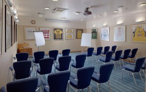 Een vergaderzaal met rijen blauwe stoelen, een whiteboard en ingelijste sporttenue aan de muren.