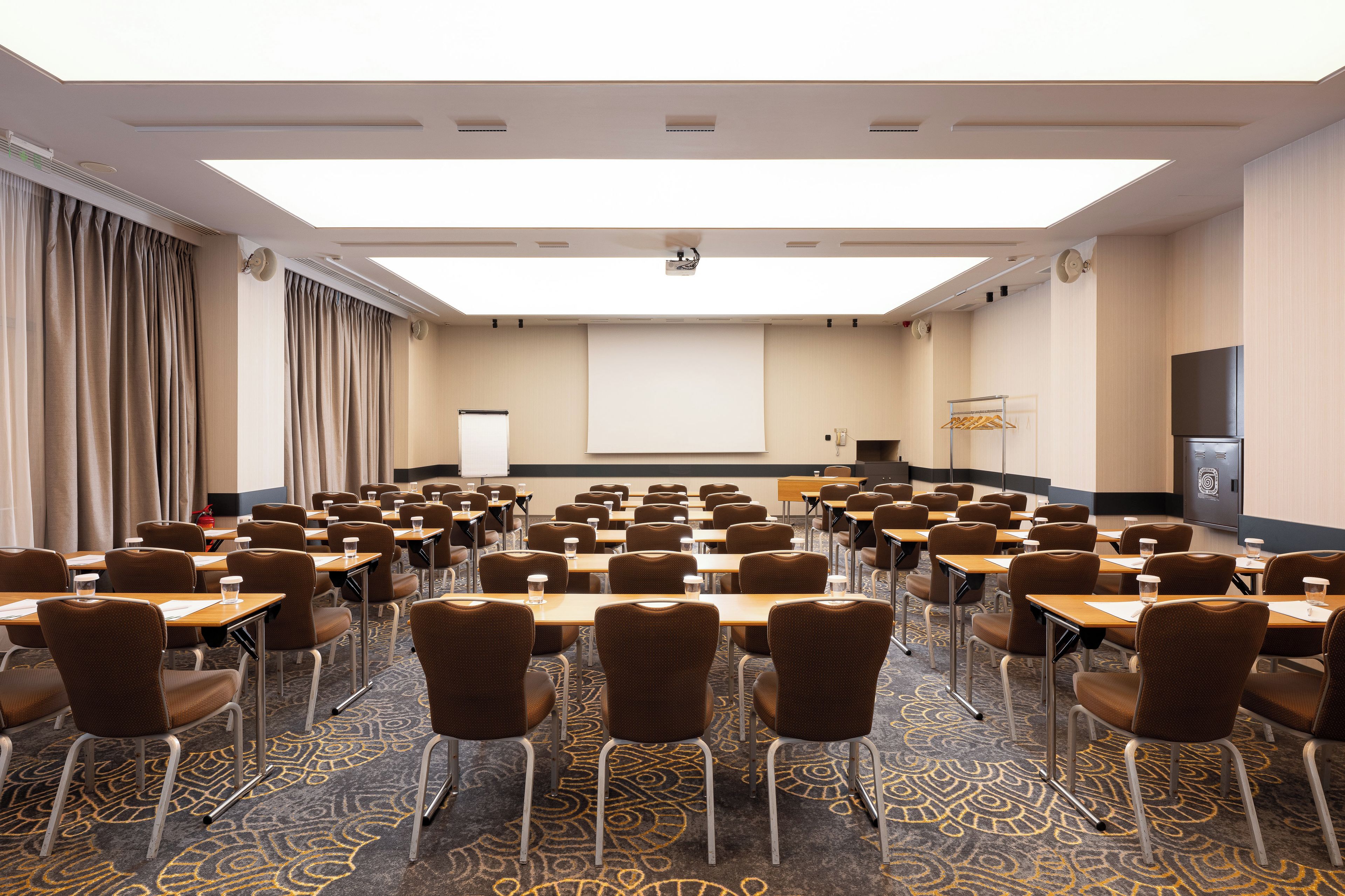 A conference room with rows of tables and chairs, a projector screen, and a large window with curtains.