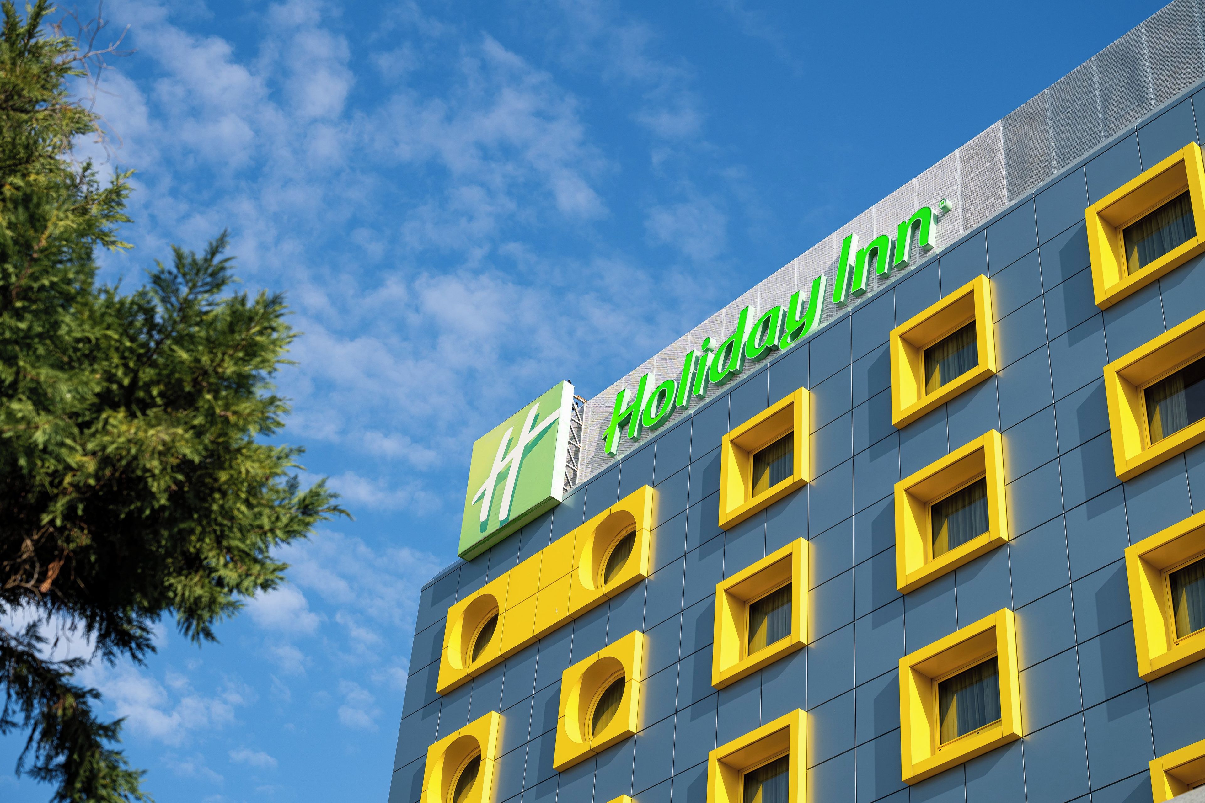 A Holiday Inn building with a clear blue sky and green foliage in the foreground.