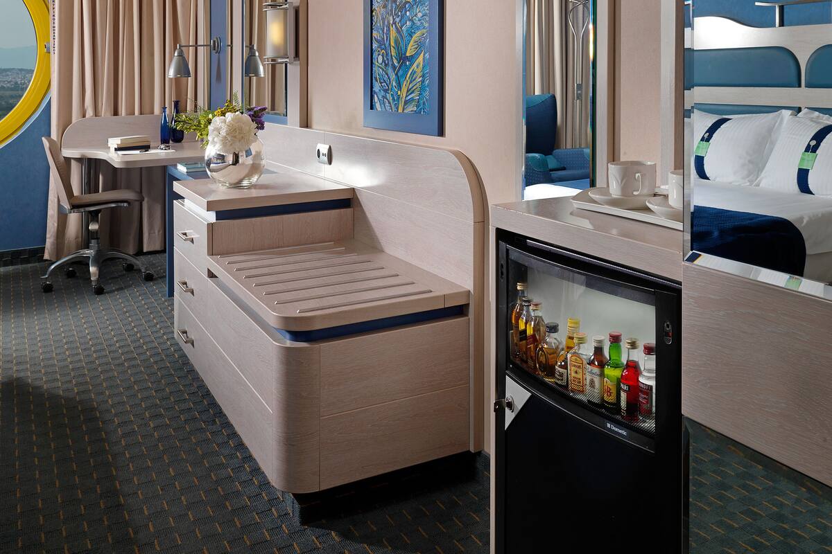 Een hotelkamer met een bureau, een minibar, een bed, een grote spiegel en een schilderij aan de muur.