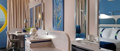 A hotel room with a desk, a minibar, a bed, a large mirror, and a painting on the wall.