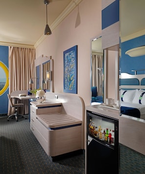 Een hotelkamer met een bureau, een minibar, een bed, een grote spiegel en een schilderij aan de muur.