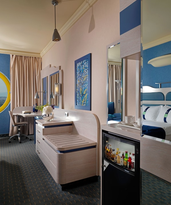 A hotel room with a desk, a minibar, a bed, a large mirror, and a painting on the wall.