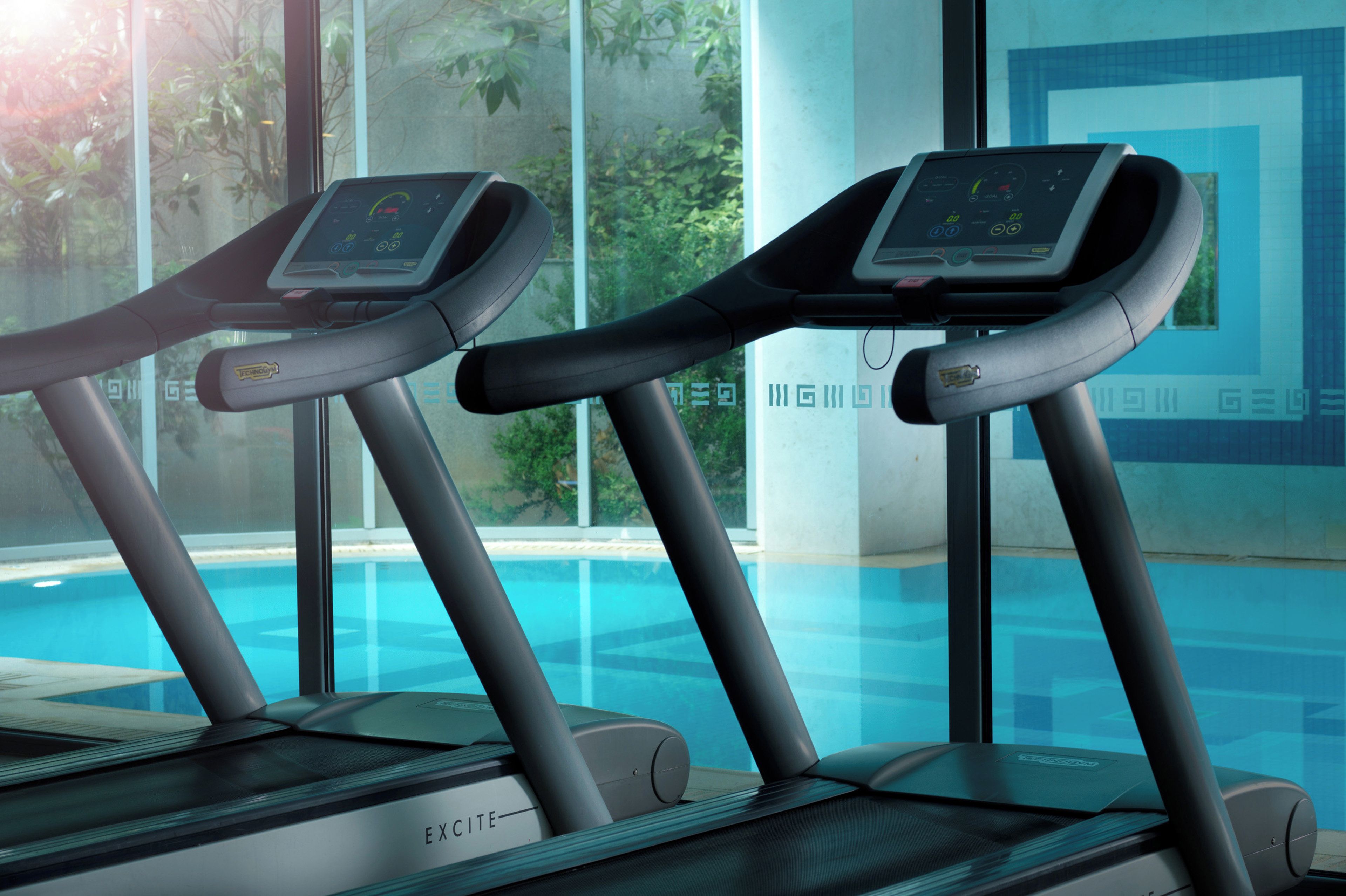Two exercise treadmills with digital displays, positioned in front of a window overlooking a swimming pool.