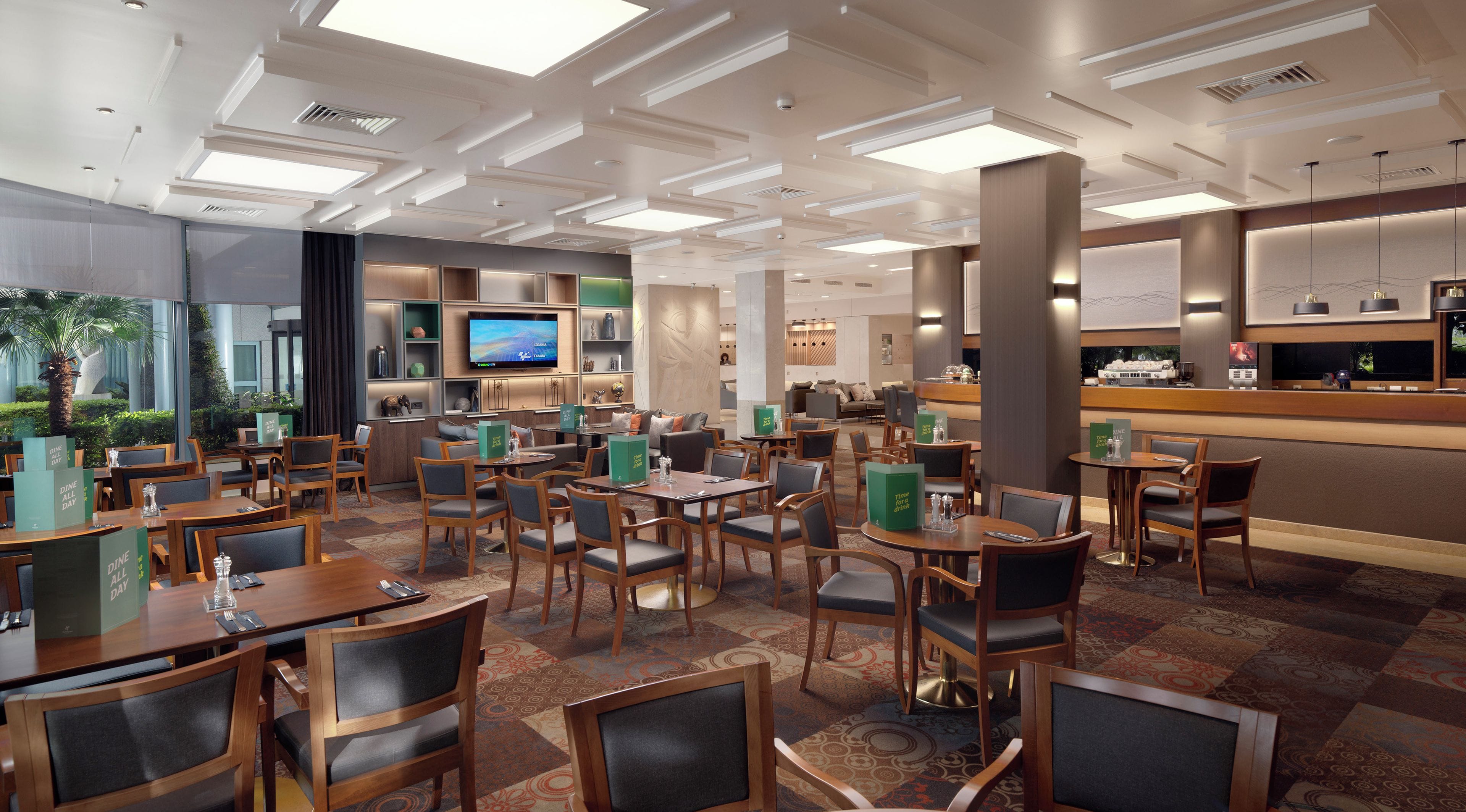 A hotel dining area with multiple tables and chairs, a television mounted on the wall, and a bar counter with a coffee machine.