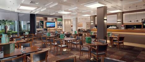 A hotel dining area with multiple tables and chairs, a television mounted on the wall, and a bar counter with a coffee machine.