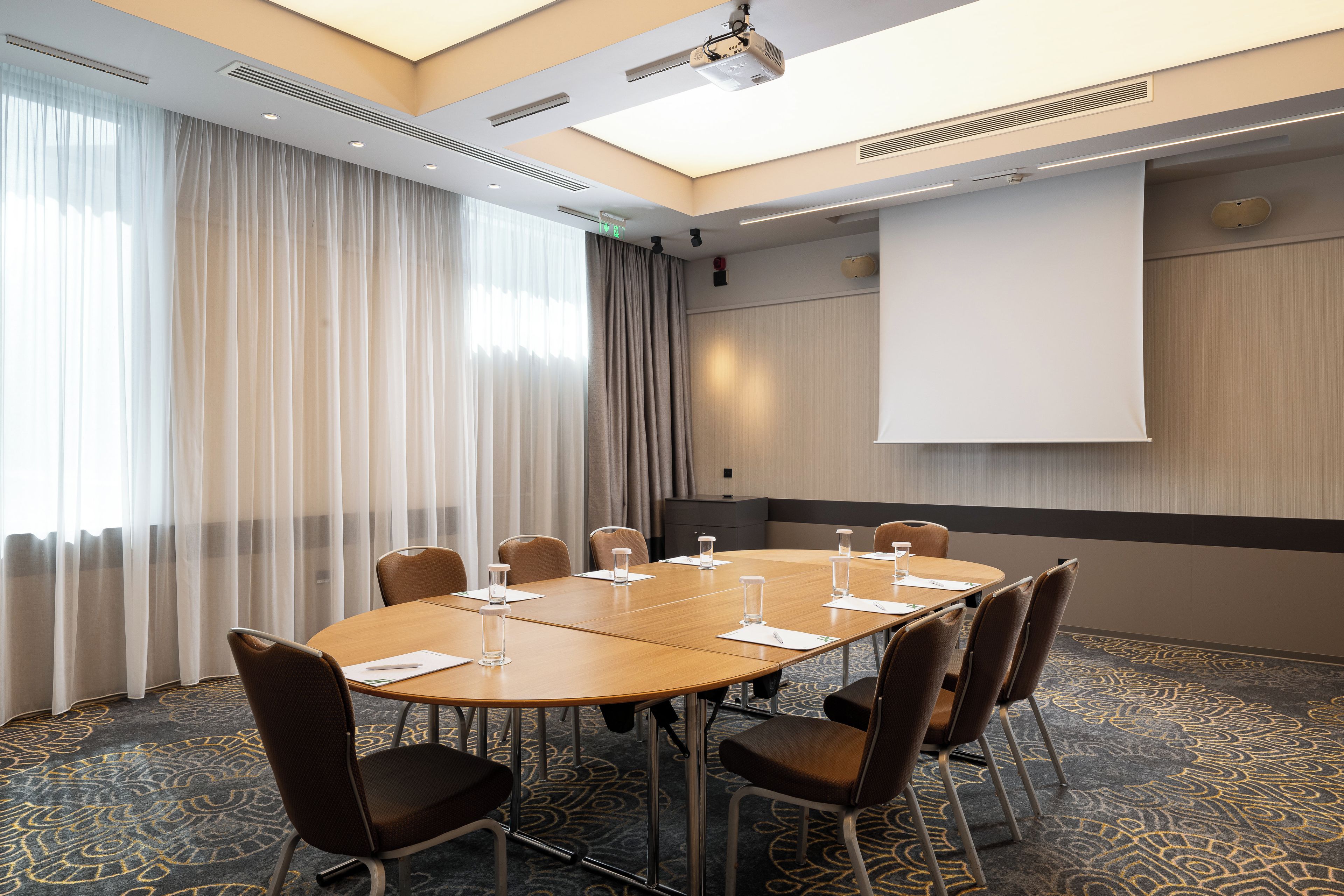 Uma sala de conferência com uma mesa grande, cadeiras, uma tela de projetor e piso carpeteado.