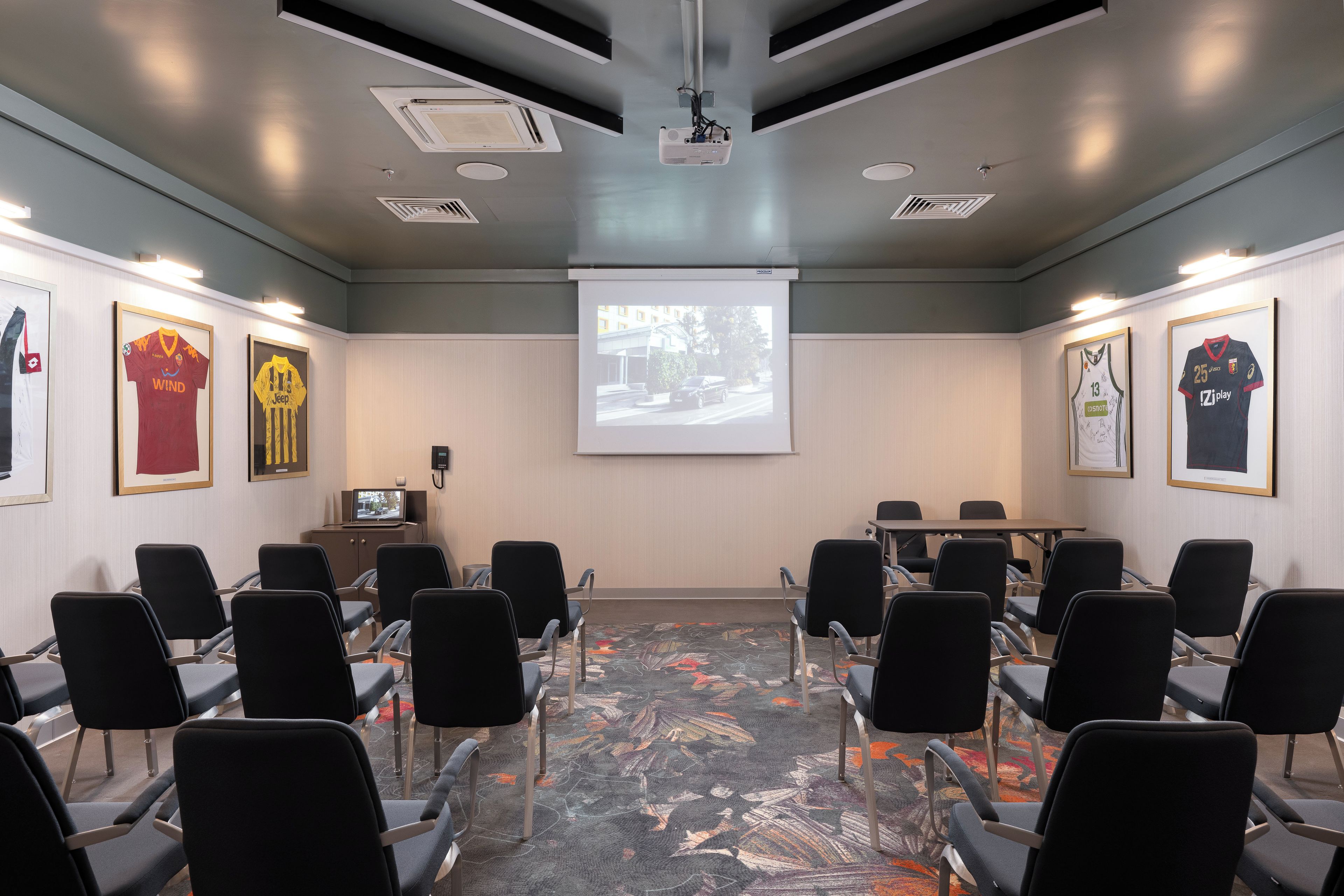 A conference room with a projector screen, black chairs, a table, and framed sports jerseys on the walls.