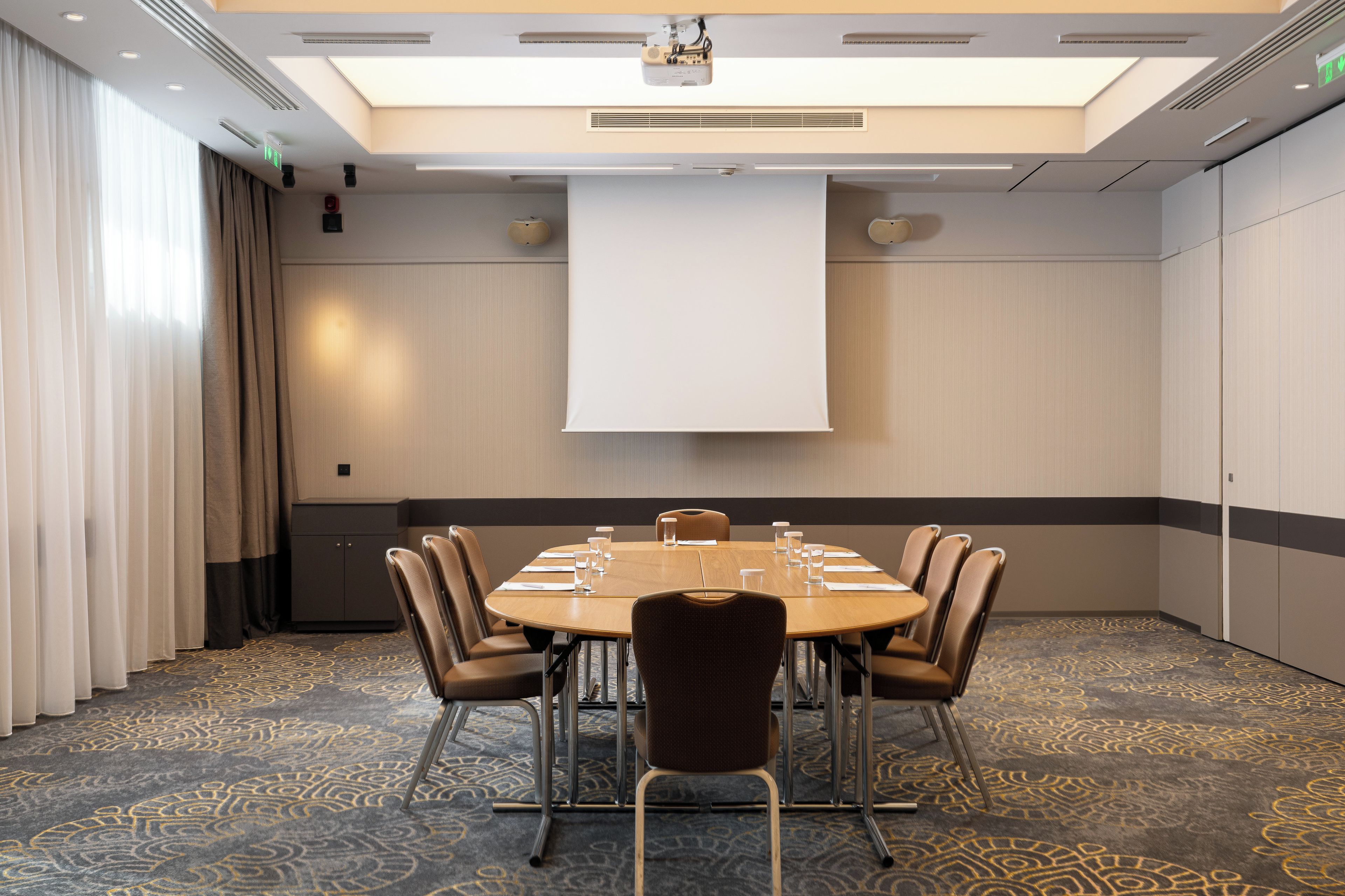 A conference room with a large table, chairs, a projector screen, and a patterned carpet.