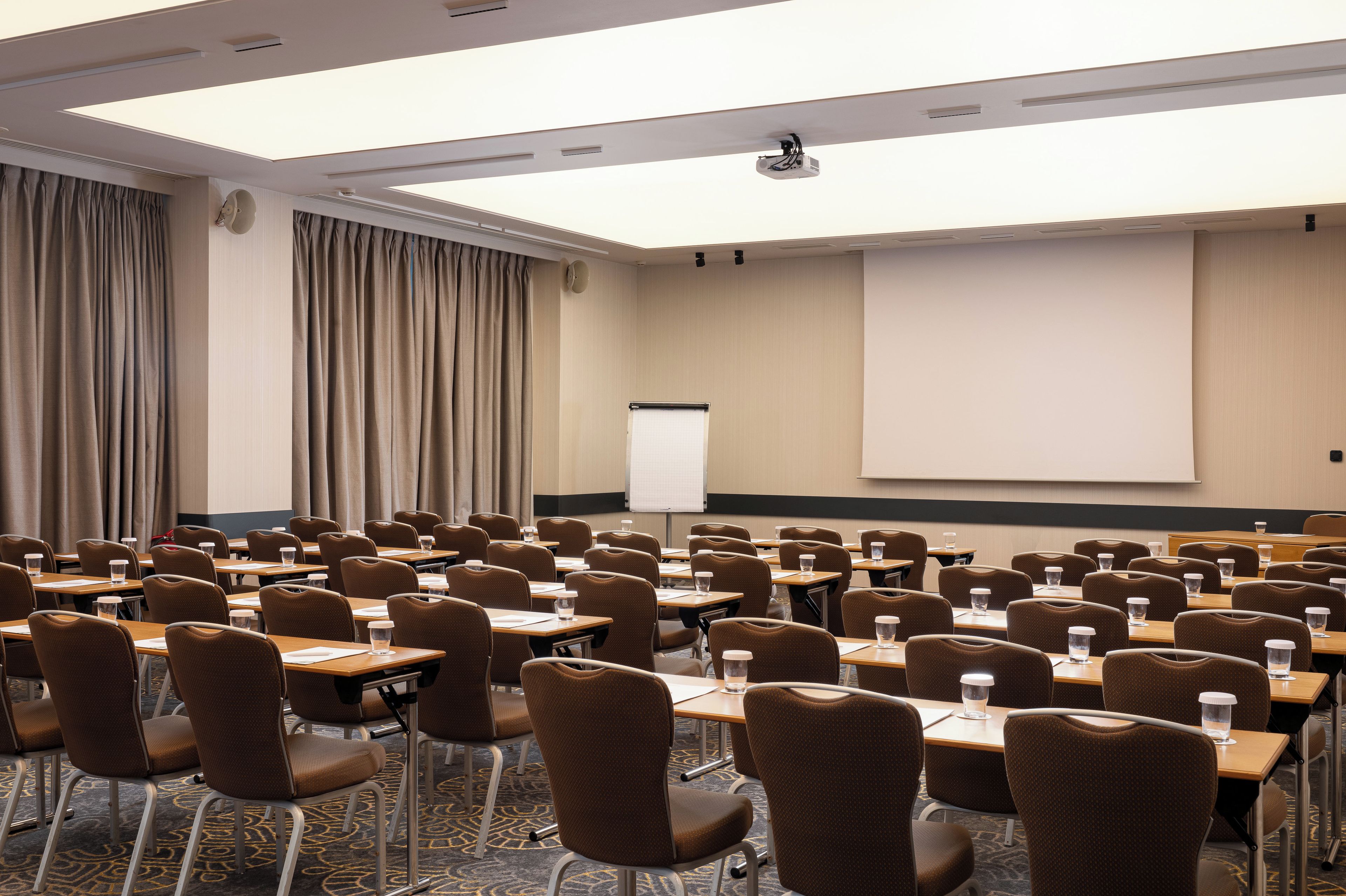 Un salón de conferencias con filas de mesas y sillas, una pantalla para proyector y una pizarra.