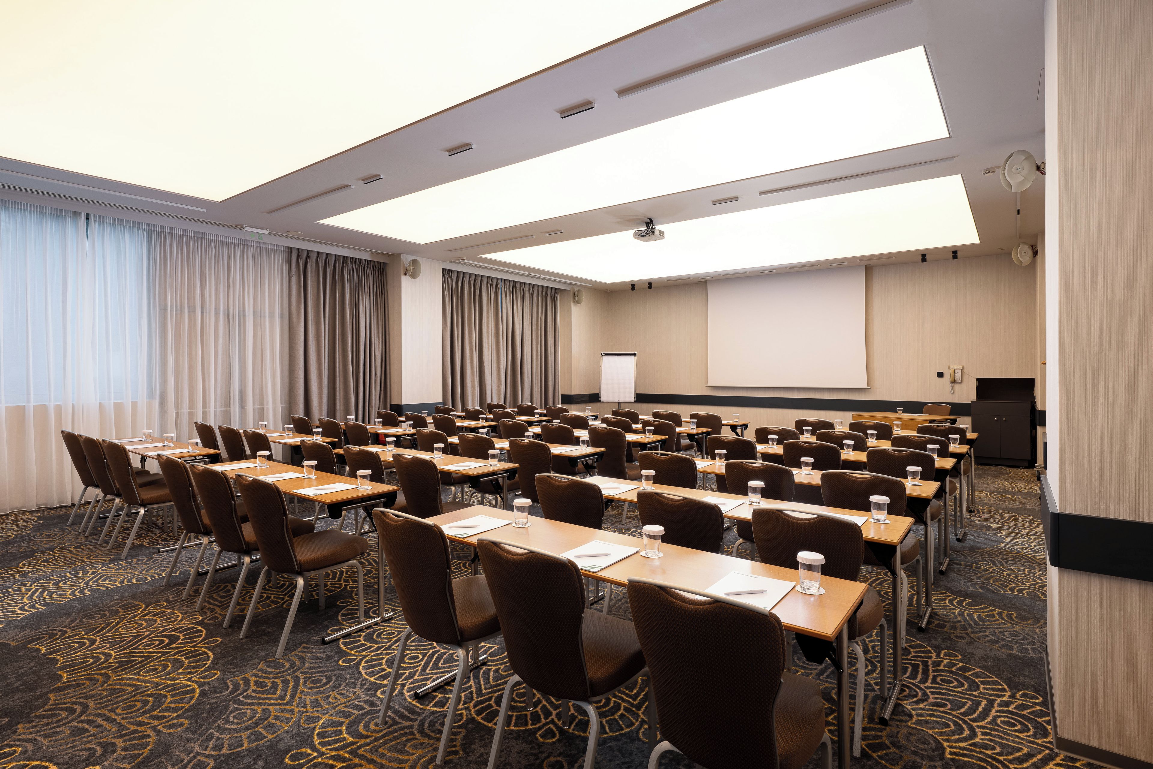 A conference room with rows of tables and chairs, a projector screen, and a large window with curtains.