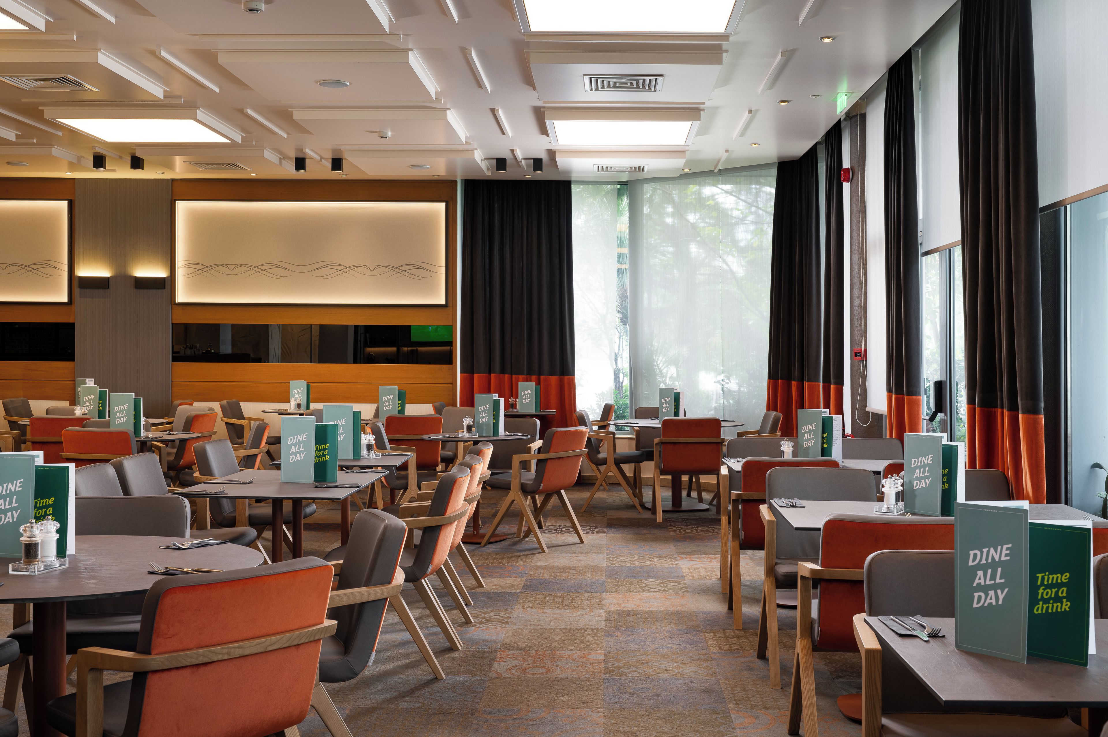 A hotel dining area with tables and chairs, 'Dine All Day' signs, and large windows with curtains.