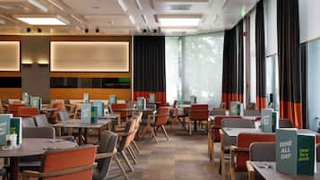 A hotel dining area with tables and chairs, 'Dine All Day' signs, and large windows with curtains.