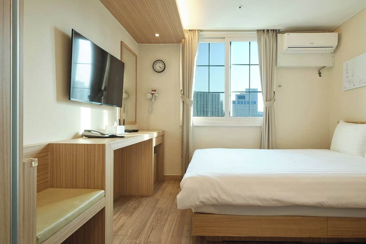 Quarto de hotel com cama, escrivaninha, televisão, relógio, ar condicionado e janela com cortinas.