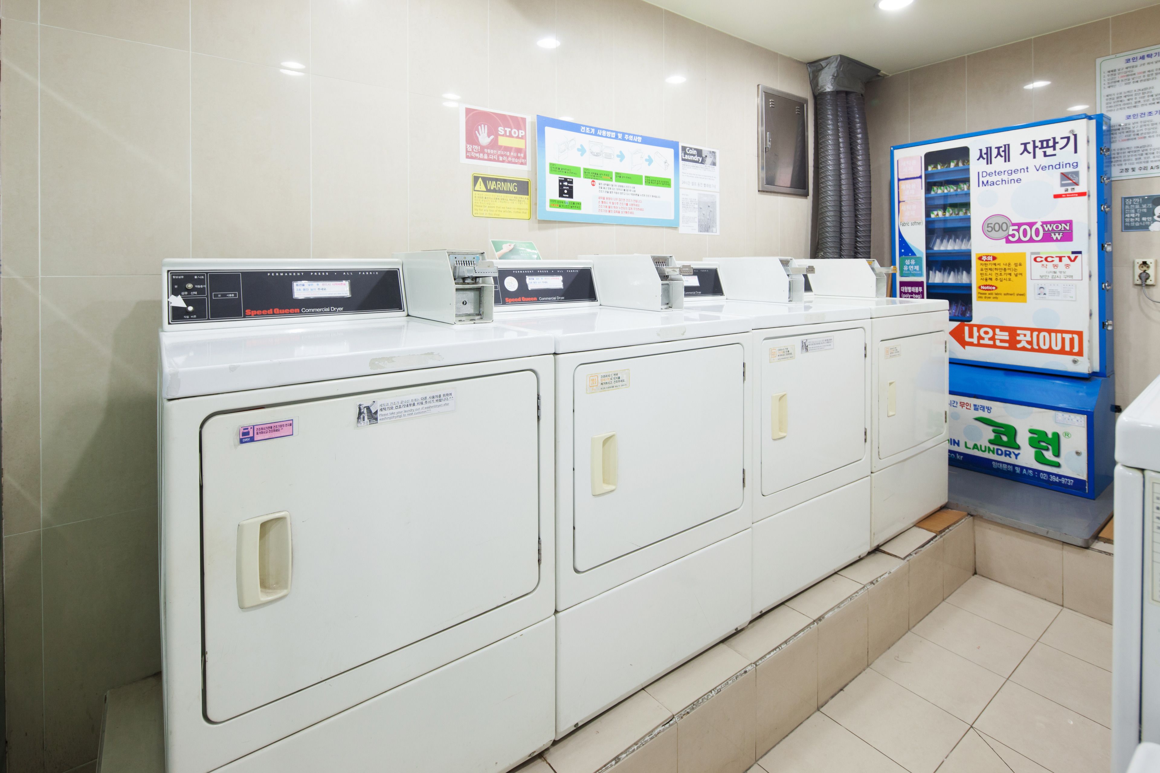 A row of white laundry machines with control panels, a detergent vending machine, and informational posters on the wall.