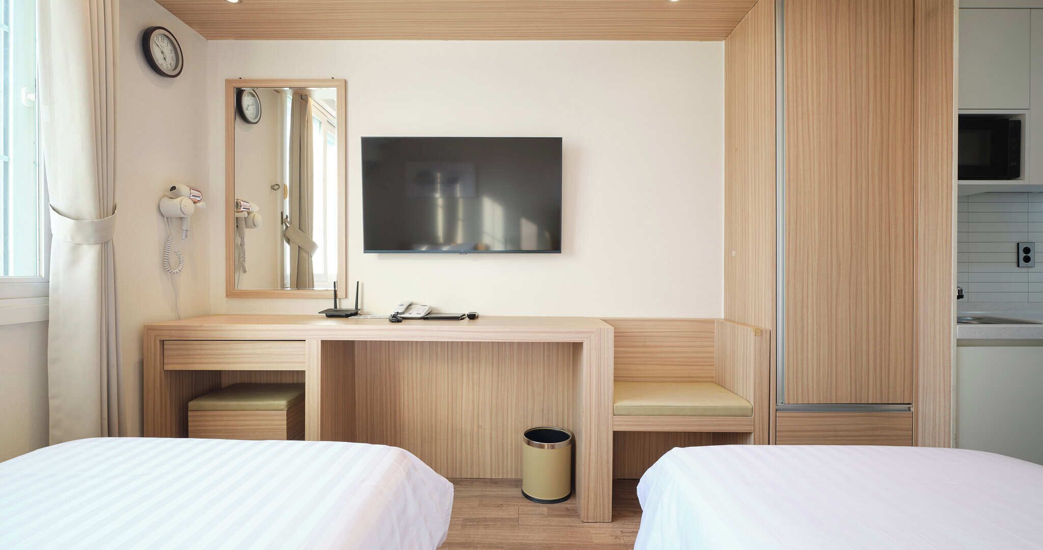 Une chambre d’hôtel équipée d’un bureau, de deux lits, d’un miroir, d’une horloge, d’une télévision et d’une kitchenette.