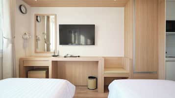 A hotel room with a desk, two beds, a mirror, a clock, a TV, and a kitchenette.