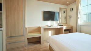 A hotel room with a bed, a desk with a chair, a TV, a mirror, a clock, and a window with a view of buildings. - Uljiro Co-Op Residence (Seoul)