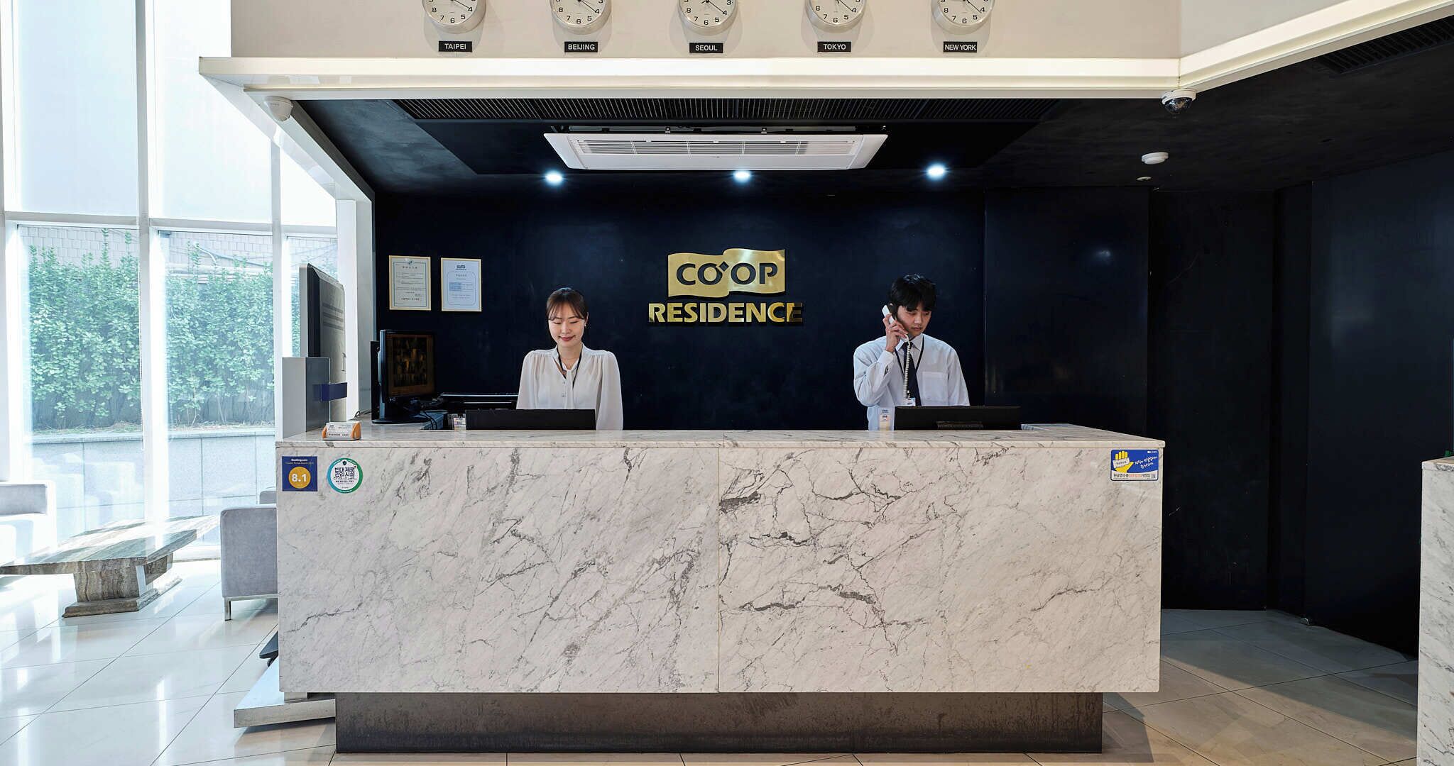 A hotel reception desk with a marble counter, clocks displaying multiple time zones, and two staff members.