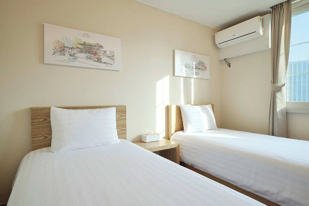 Quarto de hotel com duas camas, uma mesa de cabeceira, um aparelho de ar condicionado e duas obras de arte penduradas na parede.