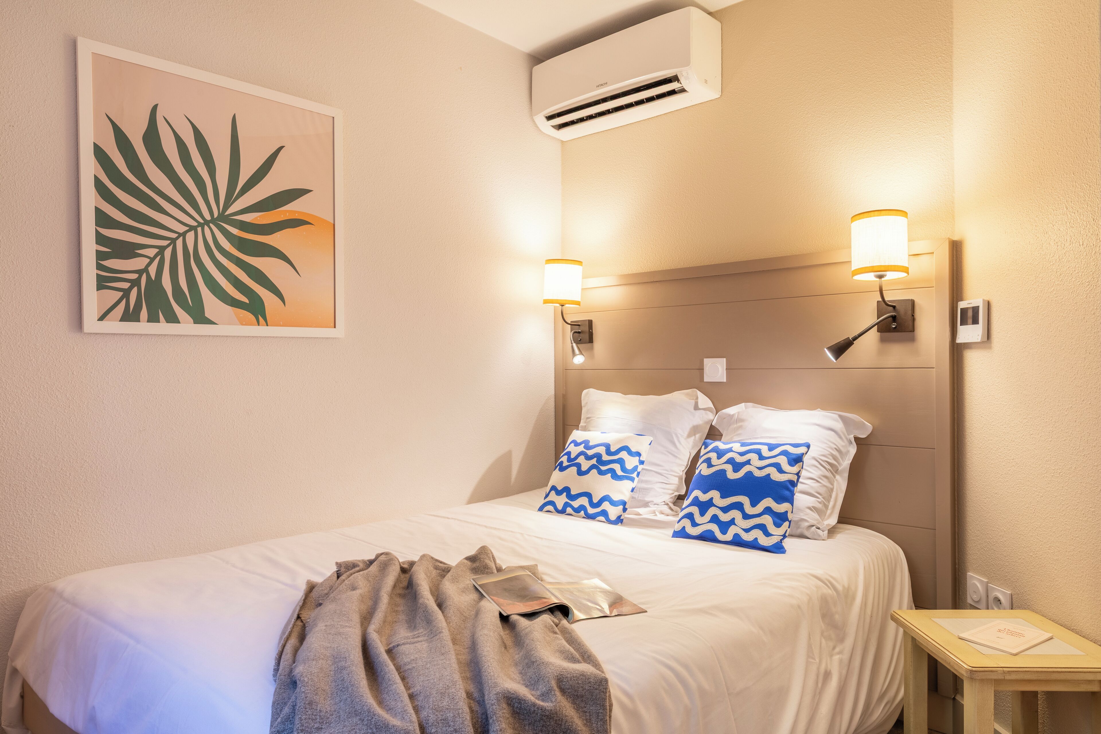 A neatly made bed with white and blue patterned pillows, a gray throw blanket, and a framed leaf artwork on the wall.