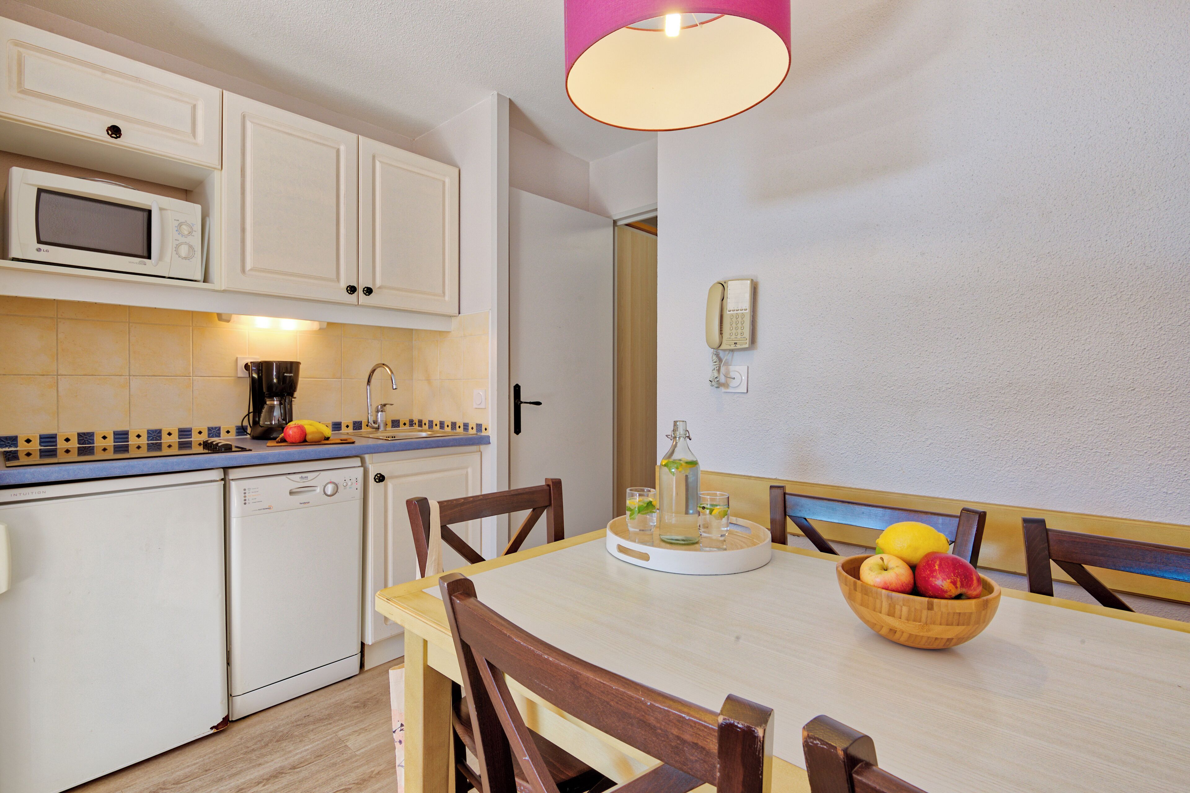 A compact kitchen with white cabinets, a microwave, and a coffee maker. There is a wooden dining table with chairs, and a fruit bowl with apples and a lemon on it.