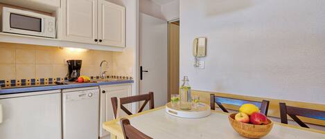 A compact kitchen with white cabinets, a microwave, and a coffee maker. There is a wooden dining table with chairs, and a fruit bowl with apples and a lemon on it.
