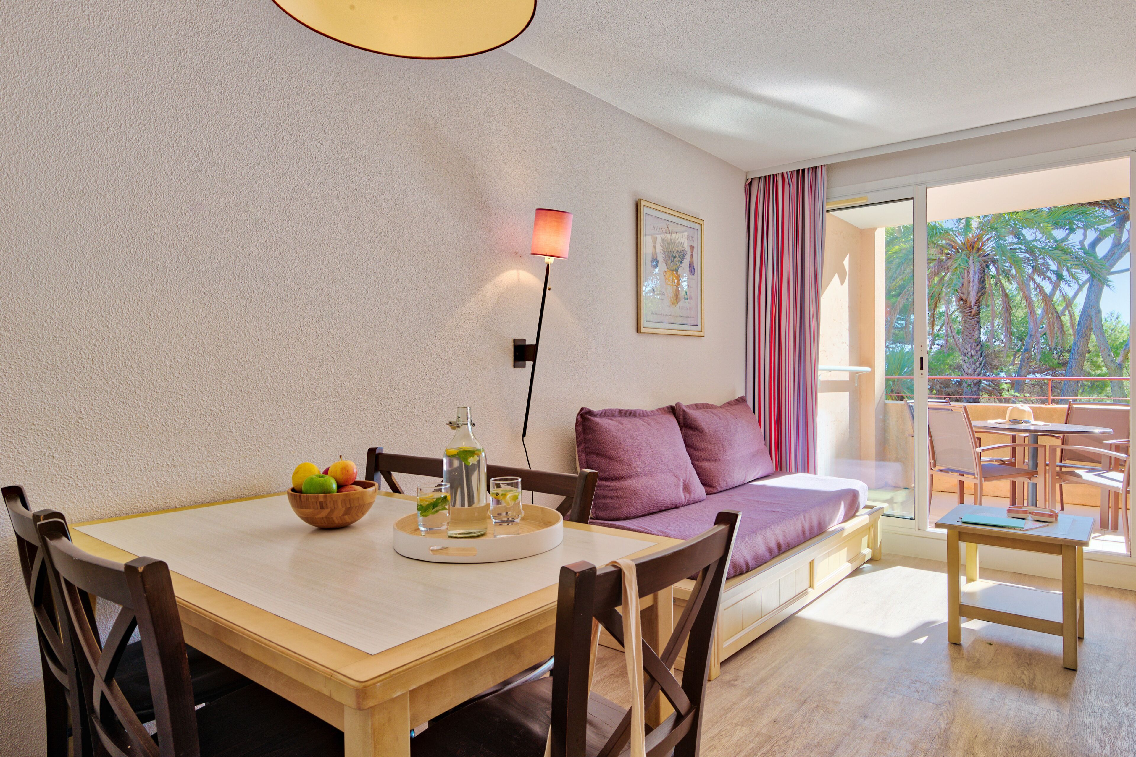 A dining area with a wooden table, chairs, a fruit bowl, and a bottle of water. There is a sofa with a cushion and a lamp on the wall.