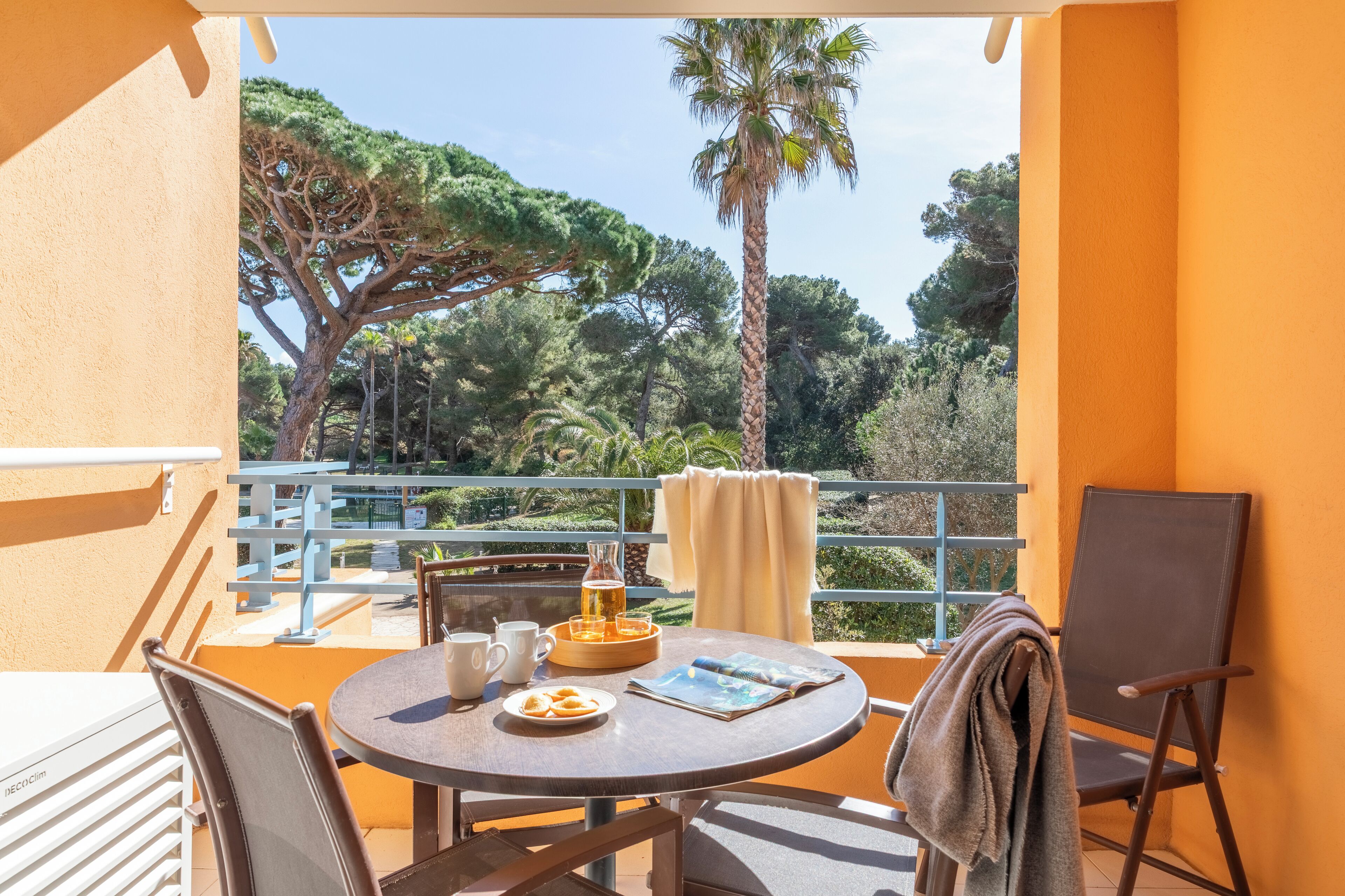 Un balcón con una mesa preparada para el desayuno, una silla y vistas a árboles y una palmera.