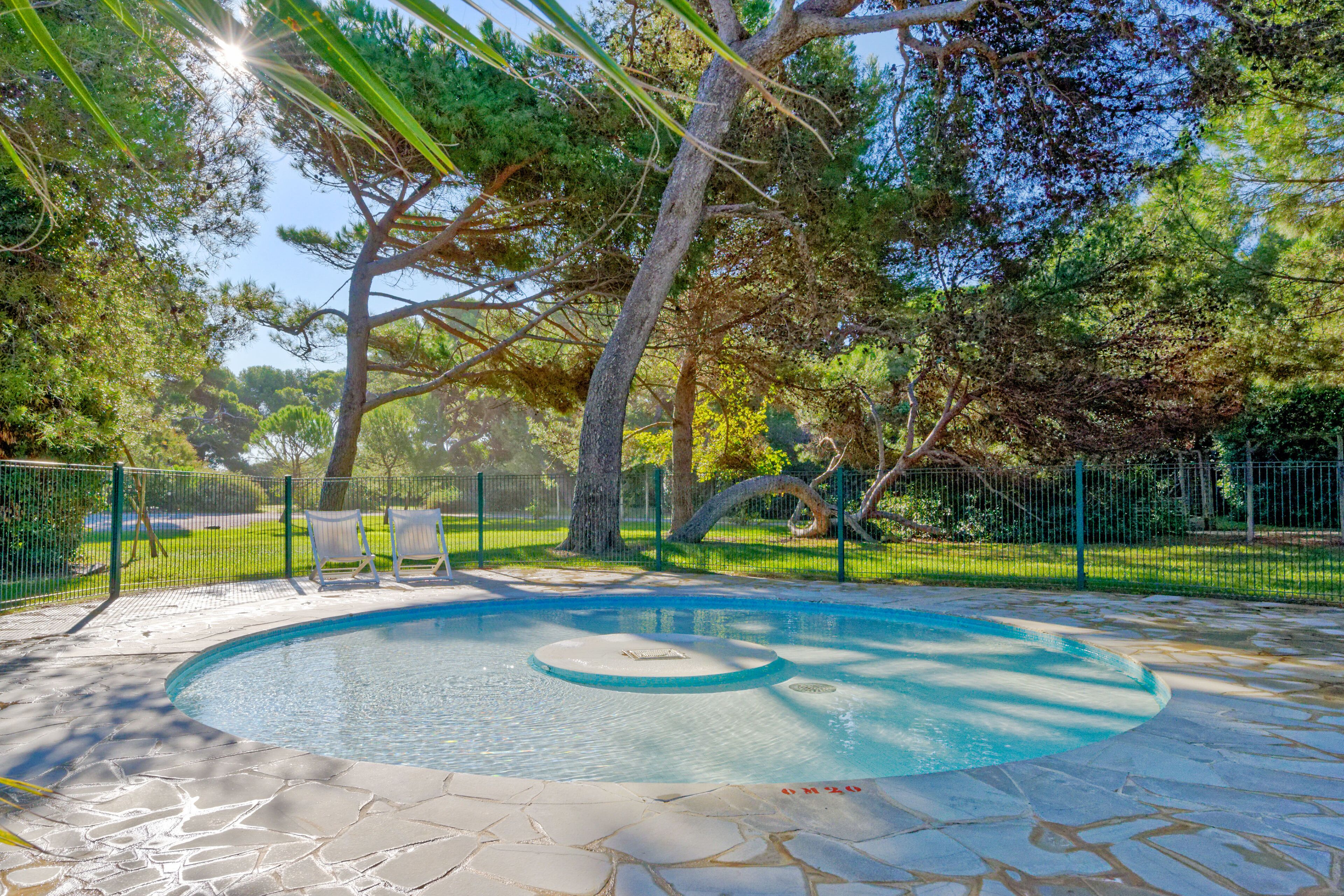 Une piscine dotée d’une plage en pierre, entourée d’une clôture et d’arbres.