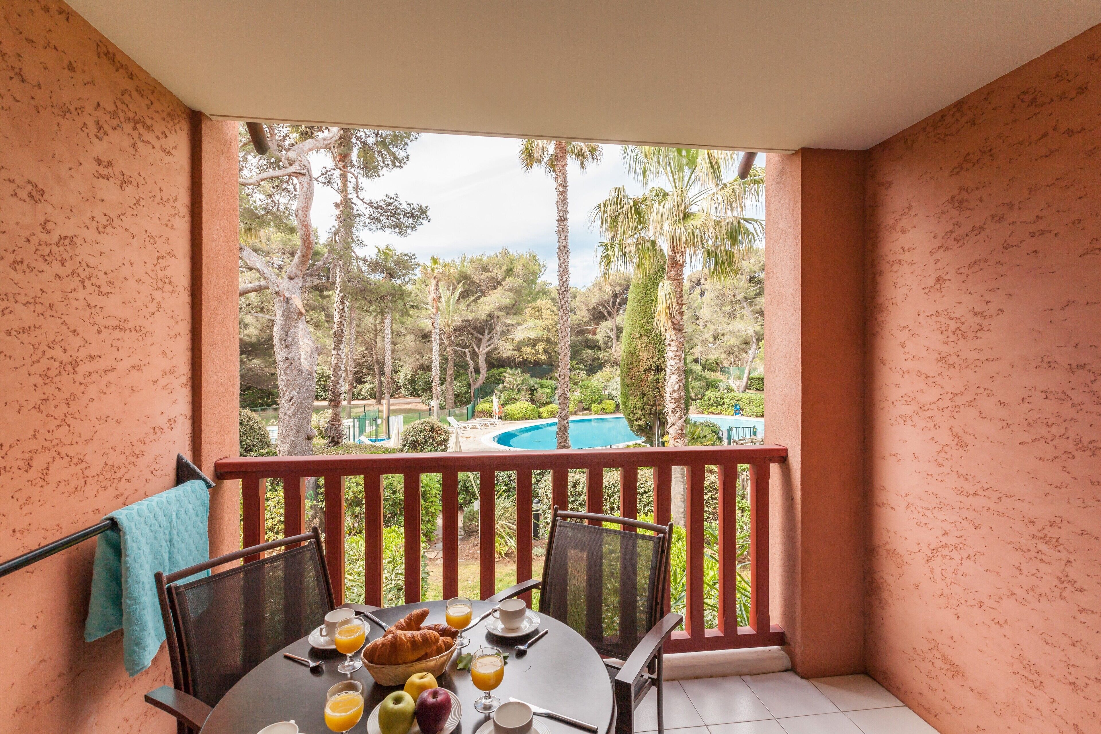 A balcony with a table set for breakfast, overlooking a swimming pool and lush greenery.