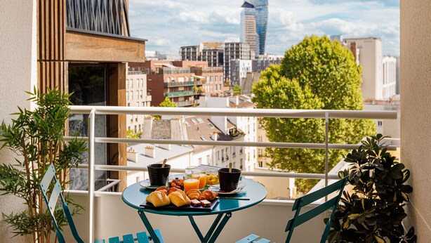 Un balcon avec une table dressée pour le petit-déjeuner, comprenant des croissants et du jus d’orange, donnant sur un paysage urbain avec des bâtiments modernes et un arbre.