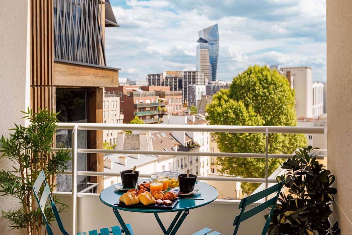 Un balcon avec une table dressée pour le petit-déjeuner, comprenant des croissants et du jus d’orange, donnant sur un paysage urbain avec des bâtiments modernes et un arbre.