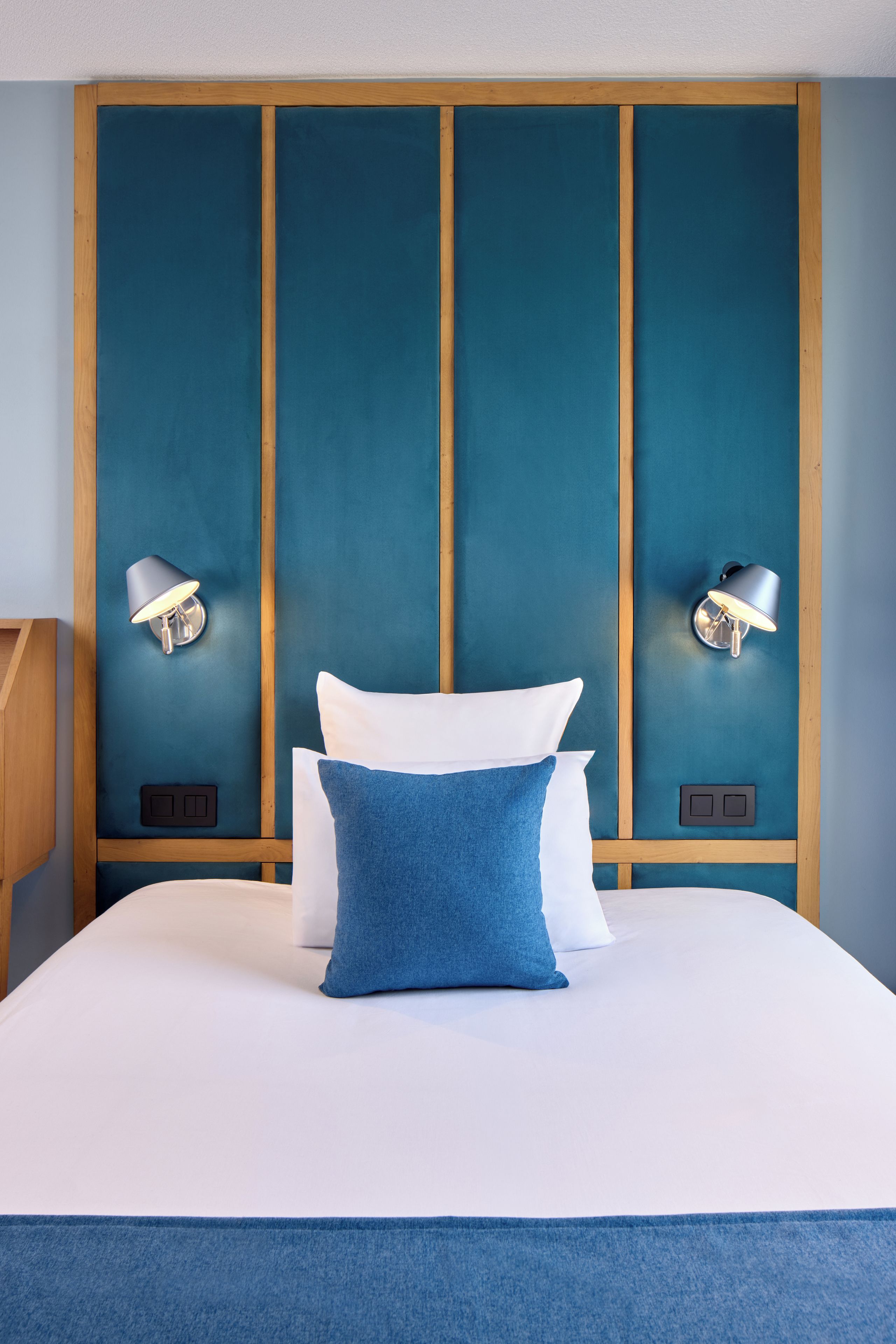 A hotel room with a large bed, two wall-mounted lamps, and a wooden headboard.