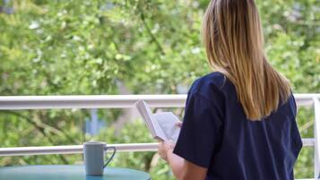A person seated at a table with a book and a mug, overlooking a balcony with greenery.