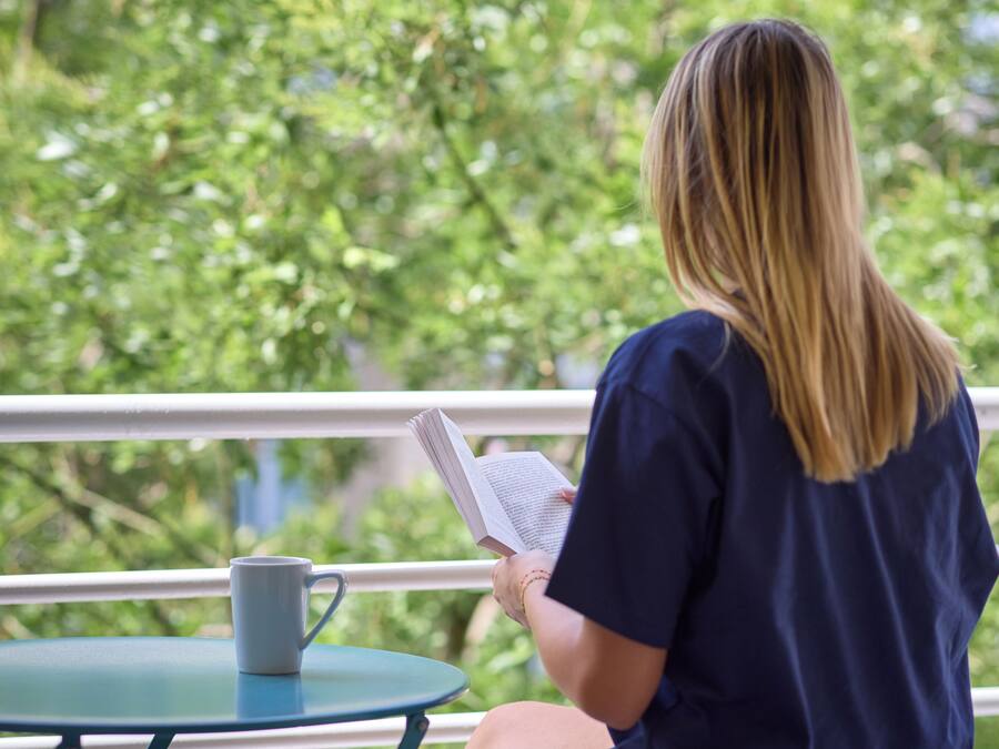 A person seated at a table with a book and a mug, overlooking a balcony with greenery.